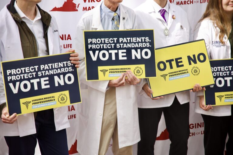 Medical professionals in white coats hold large signs reading Vote No at a protest.