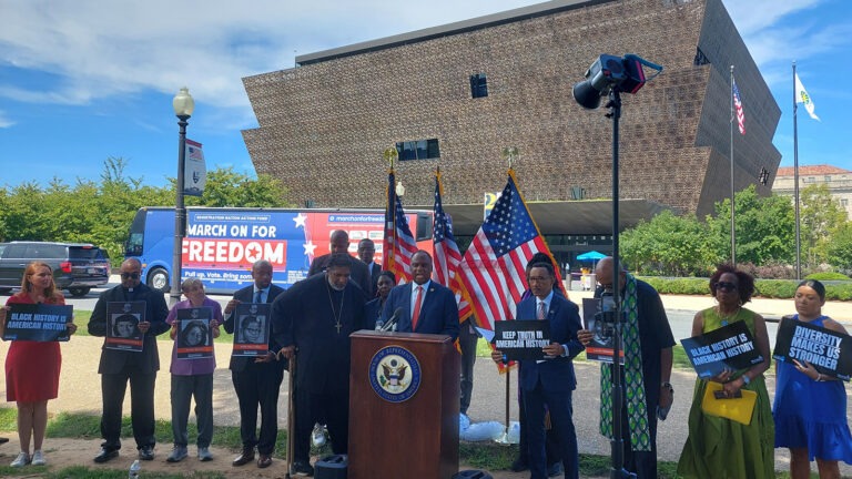 Diverse group at a podium outdoors, holding signs about Black history; U.S. flags and a 'March On For Freedom' bus behind them near a modern geometric building.