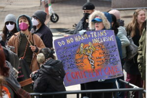 featured-image-21 - New Social Gospel Group of protesters wearing masks and winter clothing, holding a colorful sign advocating trans-inclusive healthcare.