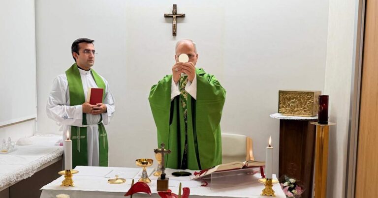 Catholic priest in green vestments elevates the Eucharist during Mass, with an assistant beside him at the altar.
