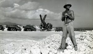 featured-image-24 - New Social Gospel Shirtless man wearing a hat and sunglasses stands with a shovel on a rocky quarry, with heavy trucks and a crane in the background.