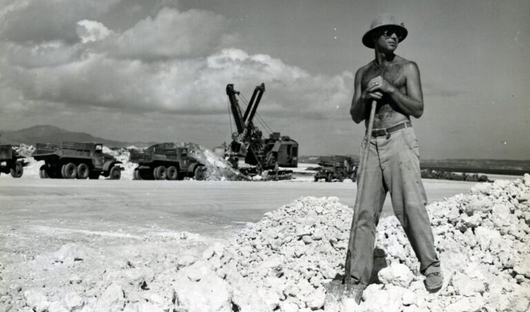 Shirtless man wearing a hat and sunglasses stands with a shovel on a rocky quarry, with heavy trucks and a crane in the background.