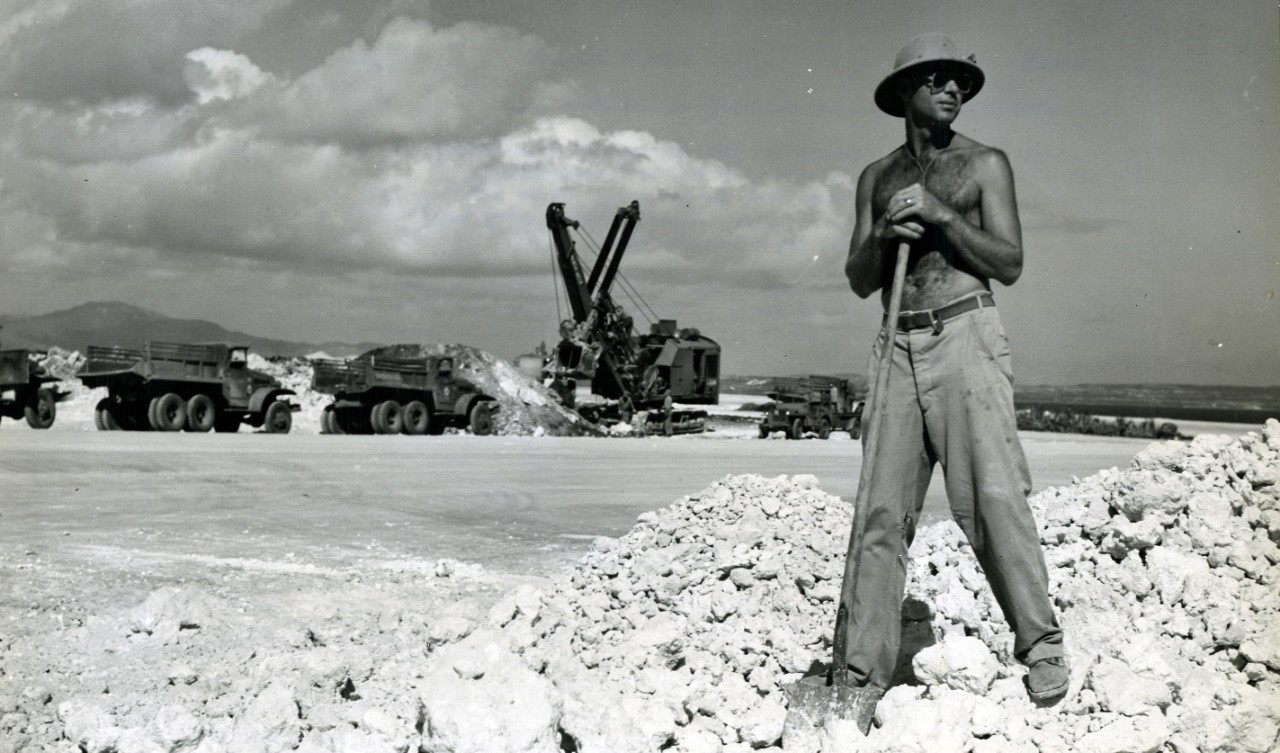 Shirtless man wearing a hat and sunglasses stands with a shovel on a rocky quarry, with heavy trucks and a crane in the background.