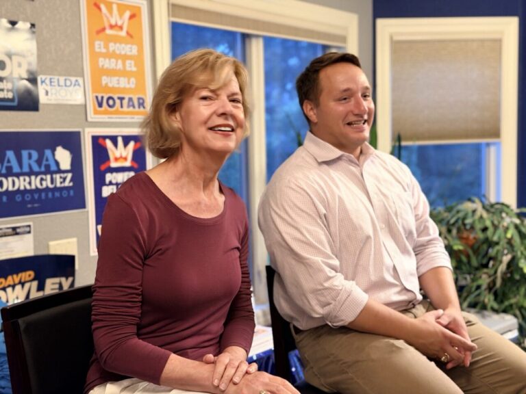 Two people sit smiling in a campaign office with posters on the wall behind them, creating a friendly scene of a political event.