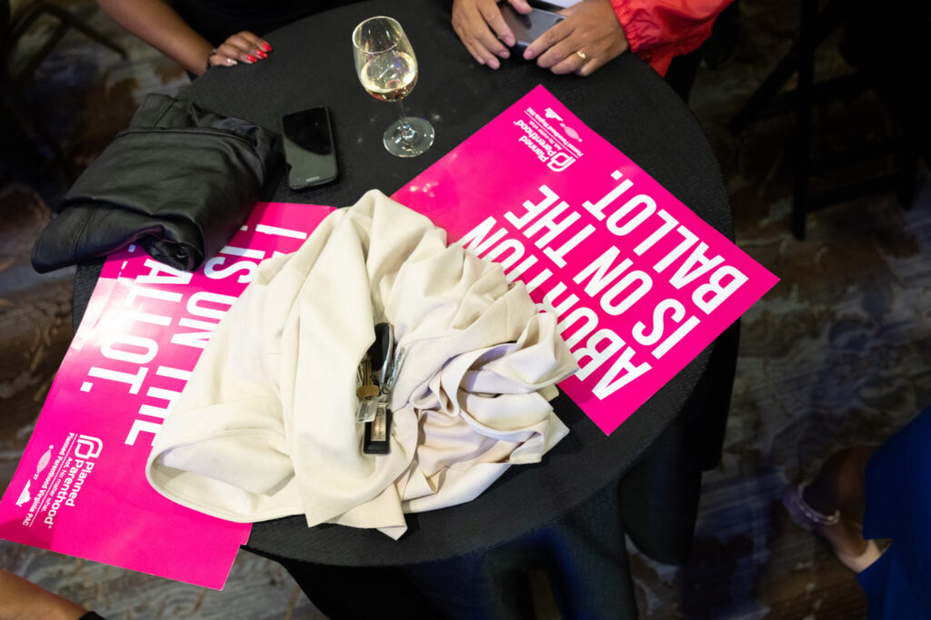featured-image-26 - New Social Gospel Top-down view of a black round table with a bright pink poster, a wine glass, a smartphone, keys, and a beige jacket laid on it left of center.