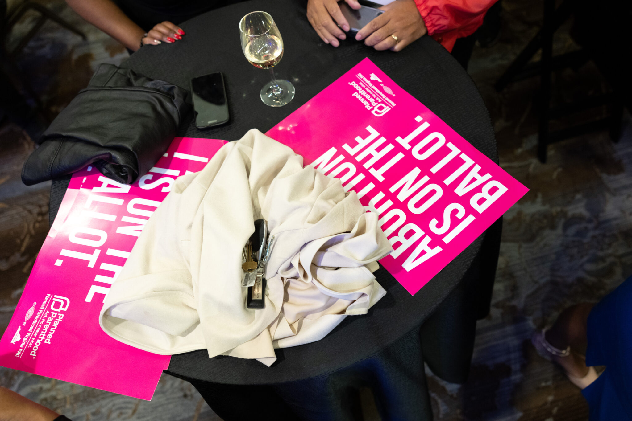 Top-down view of a black round table with a bright pink poster, a wine glass, a smartphone, keys, and a beige jacket laid on it left of center.
