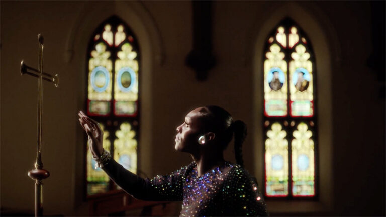 A performer in a sparkling outfit reaching toward a tall candle holder inside a church with stained-glass windows behind her.