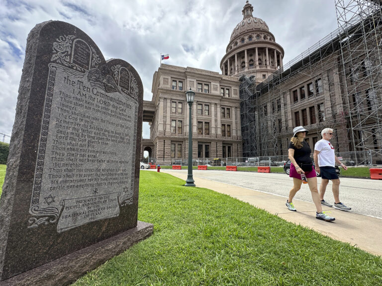 Stone Ten Commandments monument in the foreground; Texas Capitol building under scaffolding in the background with two pedestrians walking by.