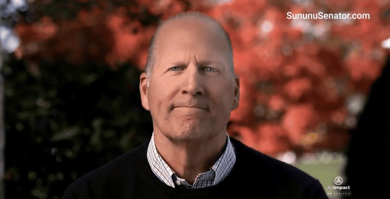 Close-up of a man speaking outdoors with red autumn leaves in the background; wearing a dark sweater over a checkered shirt; SununuSenator.com watermark on top right.