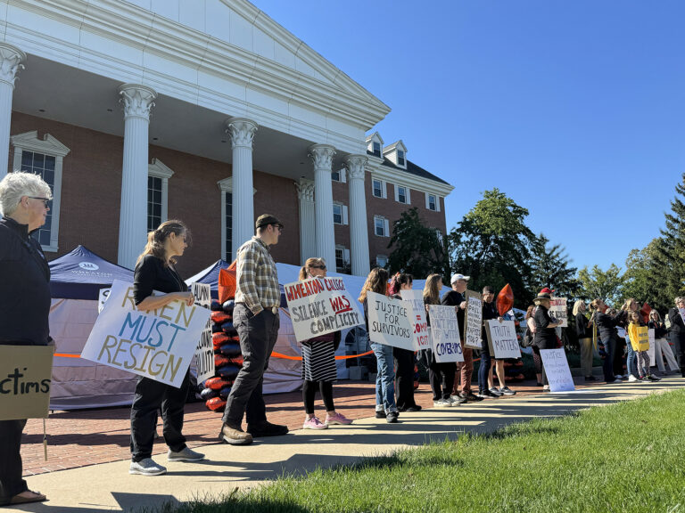 Line of protesters standing in a row outside a brick building with white columns, holding signs demanding resignation and accountability on a sunny day.