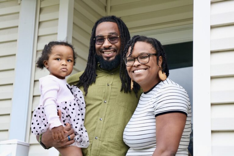 Family posing on a porch: father with glasses holding a toddler girl, mother beside them smiling.