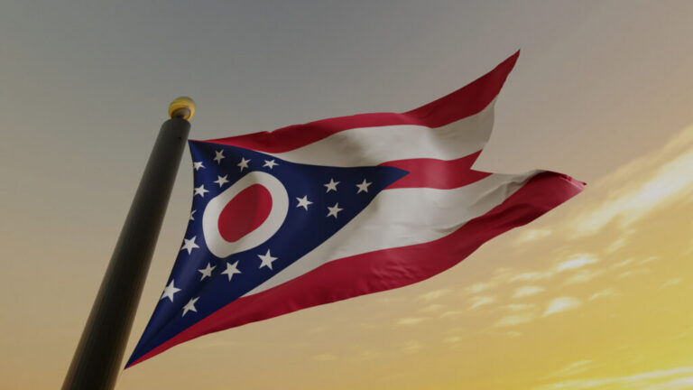 Flag of Ohio waving on a flagpole at sunset. Composed of the blue triangle with white stars and red stripes, and a red circle.