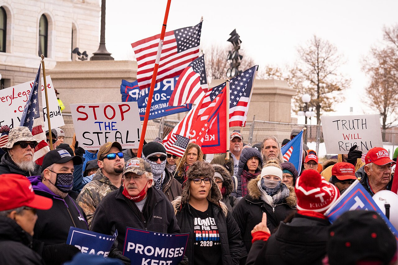 Crowd of protesters marching with American flags and signs, including 'STOP THE STEAL' and 'TRUTH WILL REBUILD' in a public square.