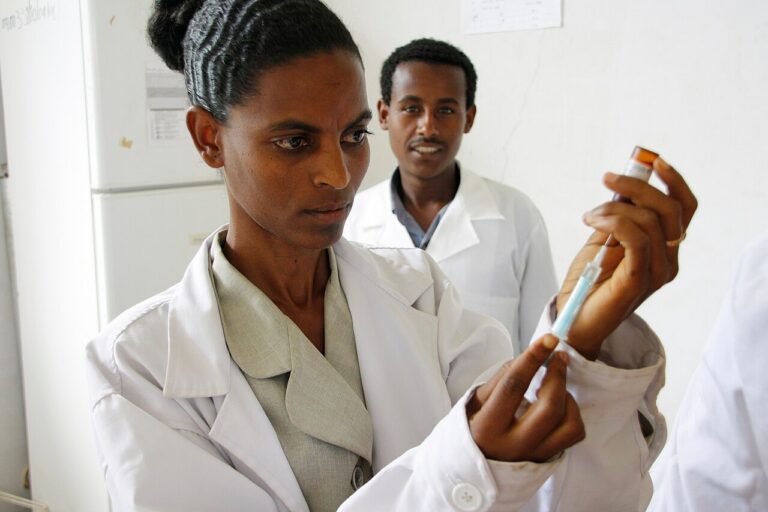 Person in a white lab coat fills a syringe from a vial as a colleague watches in a laboratory setting.