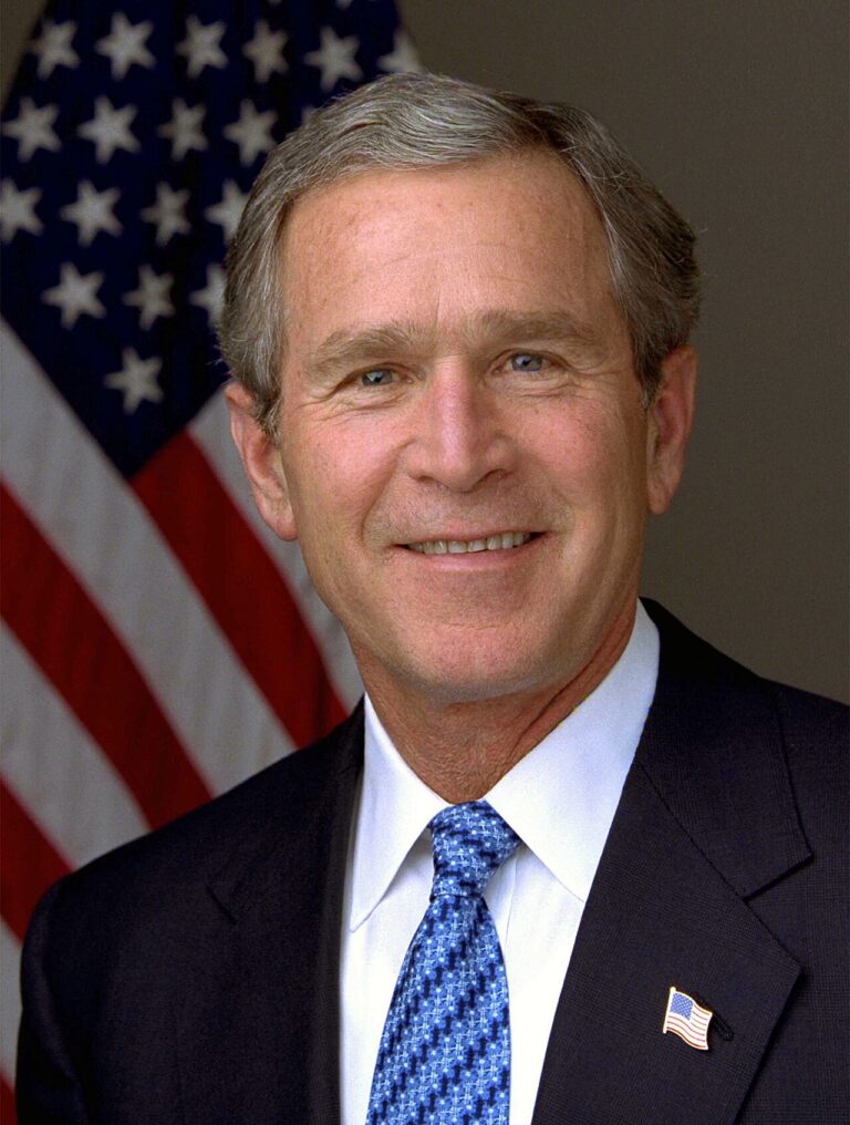 Portrait of a smiling man in a dark suit and blue patterned tie, with an American flag in the background.
